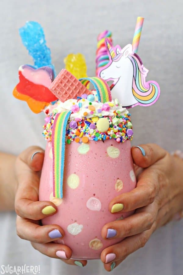 Childs hands with painted fingernails holding large pink milkshake with rainbow candy and unicorn head